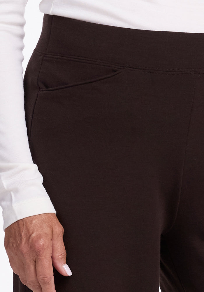 A close-up view of a person wearing black Woolx Ellie Pants with a white merino wool long-sleeved top. The image focuses on the upper thigh and waist area of the Ellie Pants, which feature a small seam detail. The person's left hand is resting by their side.