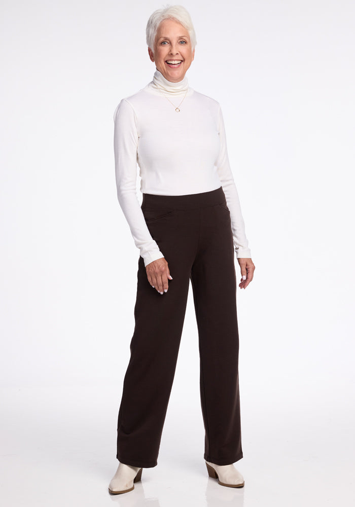 A smiling older woman with short white hair is standing against a plain background. She is wearing a long-sleeved white merino wool turtleneck shirt, Woolx black straight-leg Ellie Pants, and white ankle boots. She has her left hand resting on her thigh and appears happy and confident.