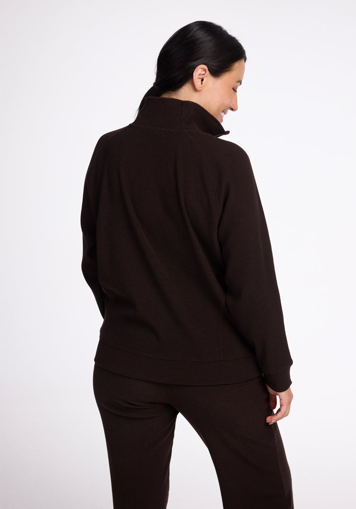 A woman with long black hair is seen from behind, wearing a dark brown Woolx Quinn 1/4 Zip sweater and matching pants. The cozy merino wool ensemble adds a touch of elegance to her fall wardrobe. She has her head turned slightly to the right, revealing a partial profile and a small smile against the plain white background. - French Roast