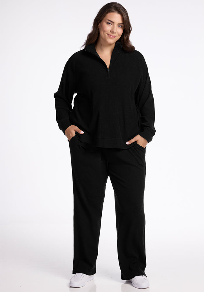 A woman stands against a plain white background, wearing a black zip-up sweater and the Woolx Luca Ribbed Pants in black. She has her hands in her pockets and is smiling, also sporting white sneakers.