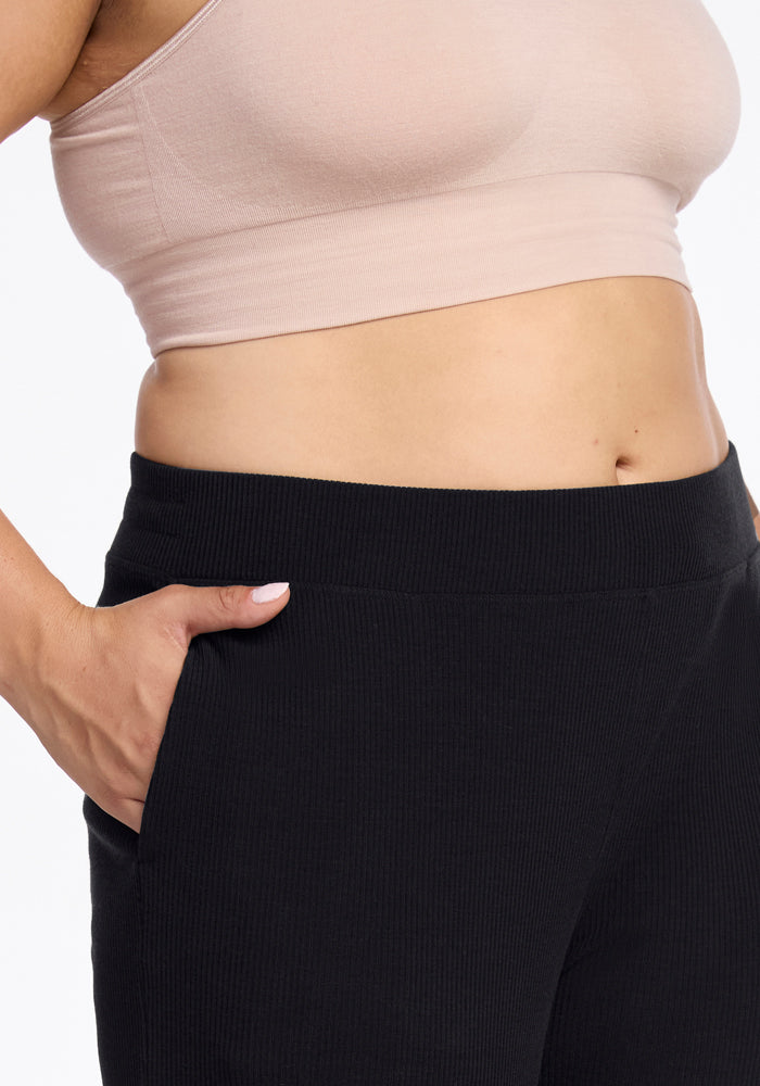 A close-up image showing the midsection of a person wearing a light beige crop top and the Luca Ribbed Pants - Black by Woolx. The person has a hand in the pocket of the pants, displaying a casual and relaxed style against a plain white background.