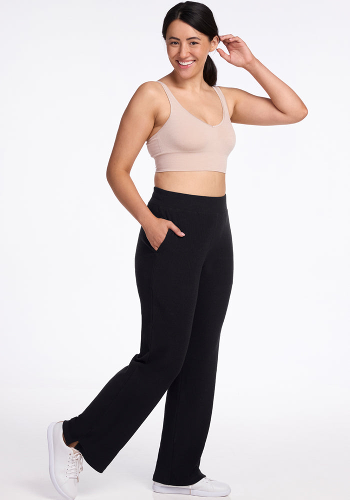 A person with dark hair is smiling and posing against a plain background. They are wearing a beige sports bra, the Luca Ribbed Pants in black from Woolx, and white sneakers. One hand is touching their hair, and the other is in their pant pocket.