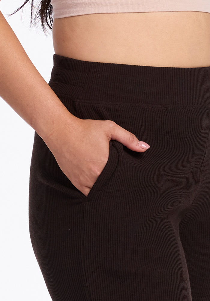 A close-up shot features a person donning the Woolx Luca Ribbed Pants in French Roast, paired with a light beige crop top. The person's right hand is tucked into the pants pocket, showcasing a peek of their upper torso. The background is simple and white, keeping the focus on the attire.