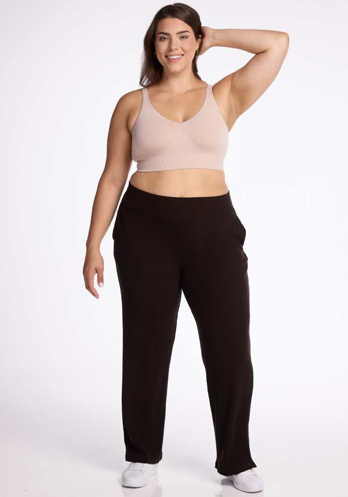 A woman with long dark hair stands smiling and posing with one hand behind her head. She is wearing a light beige sports bra, cozy **Woolx Luca Ribbed Pants in French Roast**, and white sneakers. The background is plain white, emphasizing her casual athletic attire. 