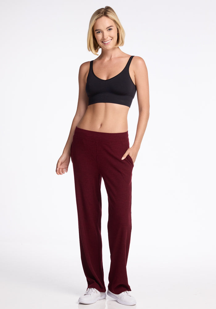 A person stands smiling in a studio setting. They have short blonde hair and are wearing a black sports bra, Luca Ribbed Pants - Cranberry Melange by Woolx, and white sneakers. One hand is in their pocket, while the other hand rests at their side. The background is plain white. 