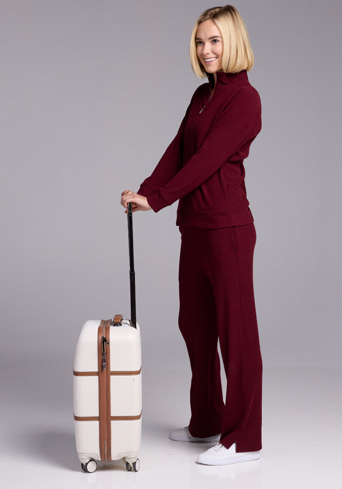 A person with blonde hair, wearing Woolx's Luca Ribbed Pants in Cranberry Melange and white sneakers, stands against a neutral background. They hold the handle of a white wheeled suitcase with brown straps, smiling and ready to travel.