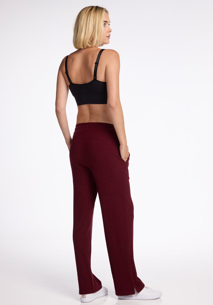 A person with blonde hair is standing and facing away from the camera. They are wearing a black sports bra and cozy Luca Ribbed Pants in Cranberry Melange from Woolx. The background is plain white.