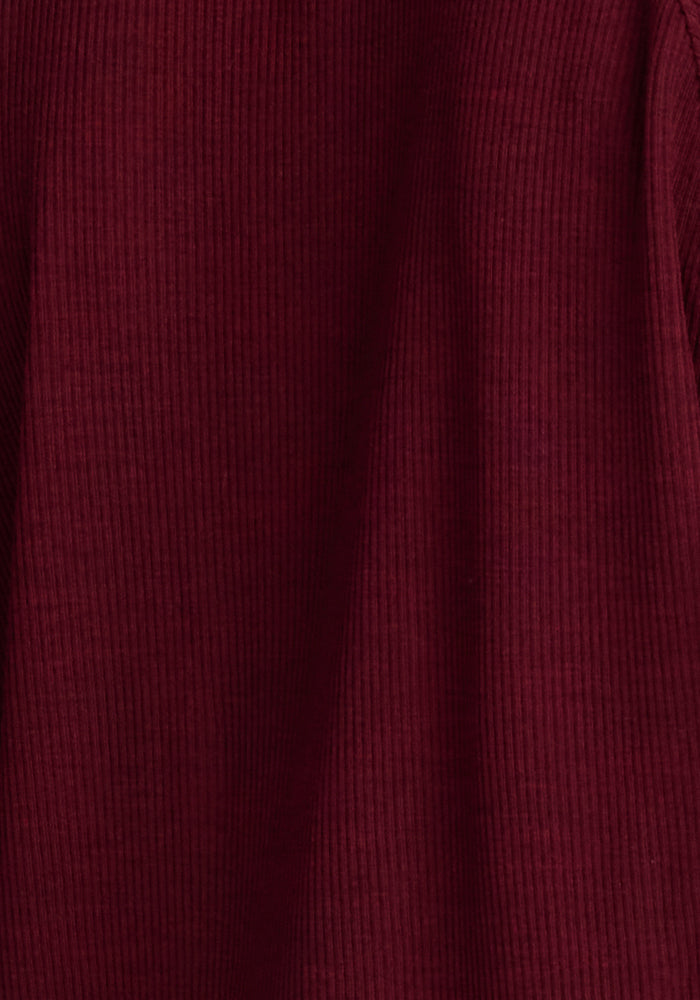 A close-up image of the Woolx Ainsley Sweater in a rich, dark red color. The Merino wool material appears to be ribbed, with vertical lines running parallel to one another, giving it a slightly ridged appearance. The lighting enhances the depth and texture of this versatile style.