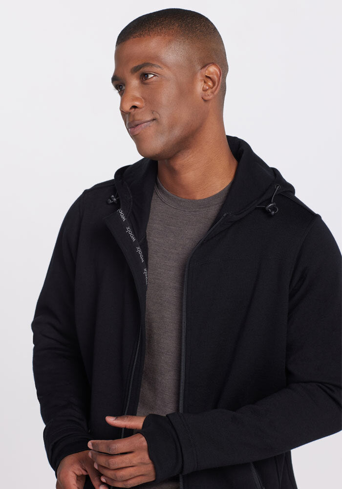 A person wearing a black, ultra-heavyweight Woolx Grizzly Sweatshirt over a grey shirt looks to the side with a slight smile. The background is plain and light-colored.