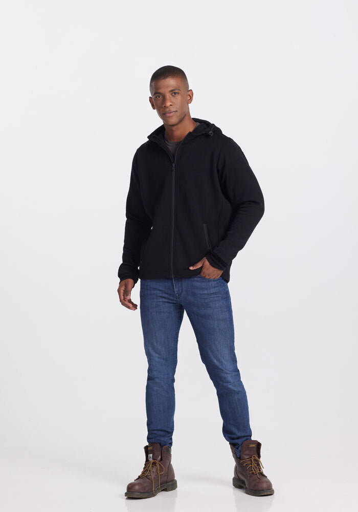 A person stands against a plain white background, wearing a Woolx Grizzly Sweatshirt in black, blue jeans, and brown boots. They have a short haircut and are casually posing with one hand in their pocket and the other hanging by their side.