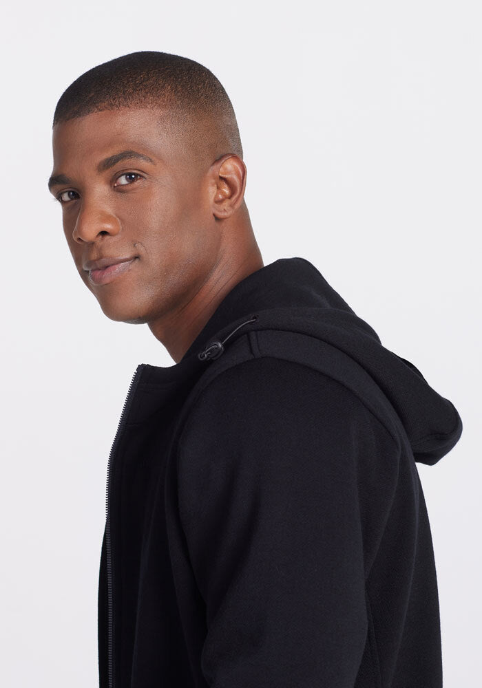 A person with short hair smiles slightly, looking over their shoulder. They are wearing a Woolx Grizzly Sweatshirt - Black with the hood down against a plain background.