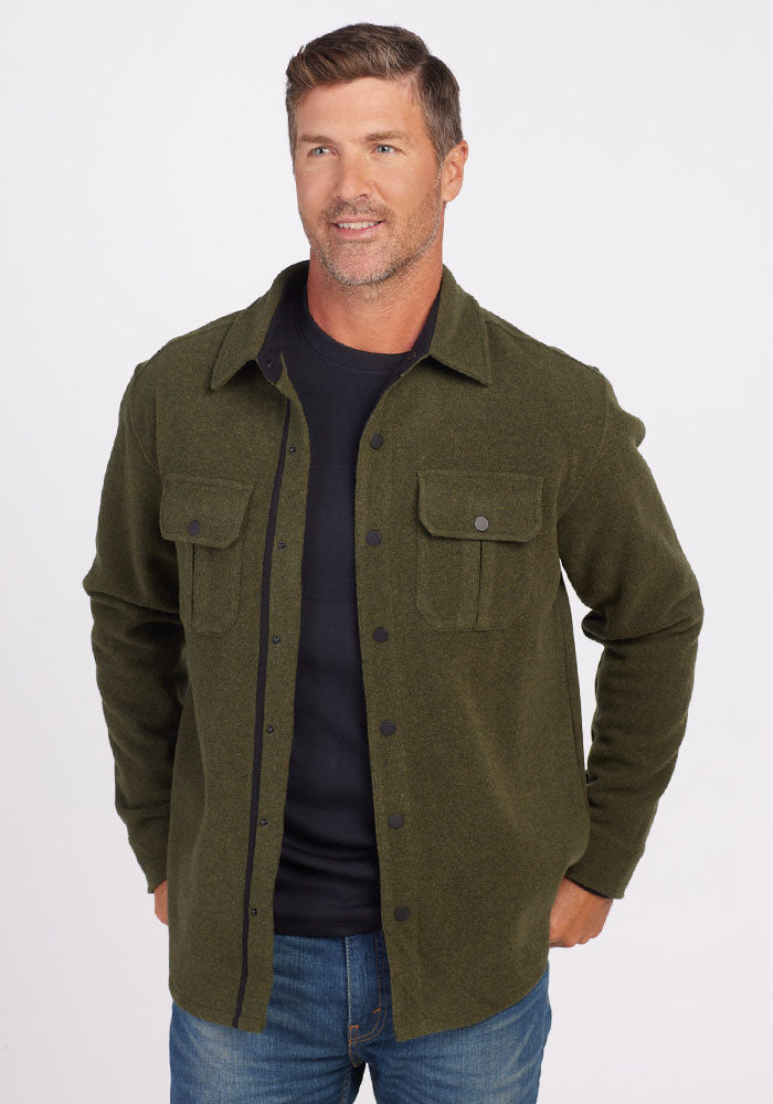 A man with short brown hair wears the Woolx Wilder Shirt Jac in Dark Forest, made from ultra-functional Merino wool, over a black shirt and blue jeans. He stands against a plain white background with his hands in his pockets, looking slightly to the side and smiling.