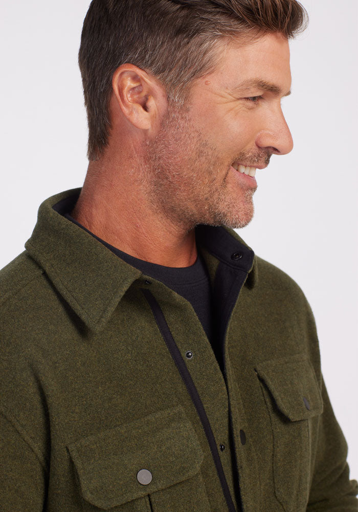 A man with short brown hair is smiling and looking to the side. He is wearing the Wilder Shirt Jac in Dark Forest over a black ultra-functional T-shirt from Woolx. The background is plain white.