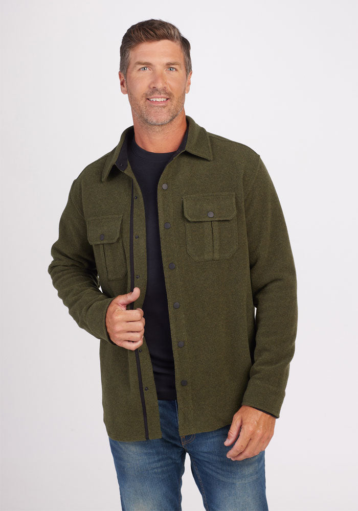 A man with short brown hair and a beard smiles while wearing the Wilder Shirt Jac by Woolx in Dark Forest over a black t-shirt. Made of ultra-functional Merino wool, this button-up shirt jacket features two chest pockets and is paired with blue jeans. The background is plain white. 