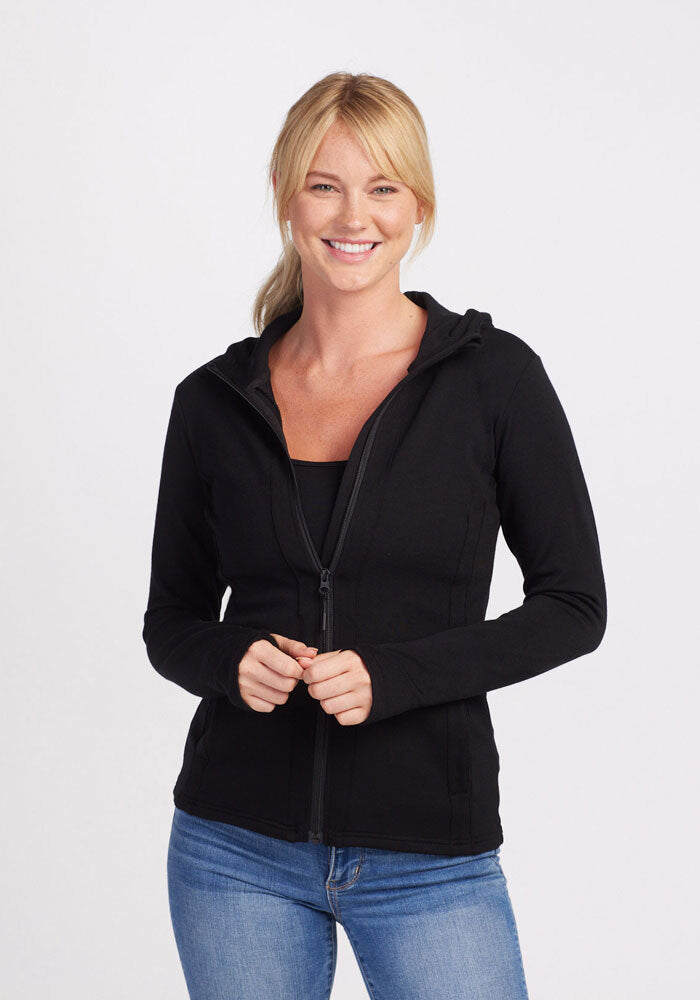 A person with blonde hair is smiling and wearing a Woolx Zoey Zip Up Hoodie in black made from DuraLite® Fabric over a dark top and blue jeans. The background is plain white.