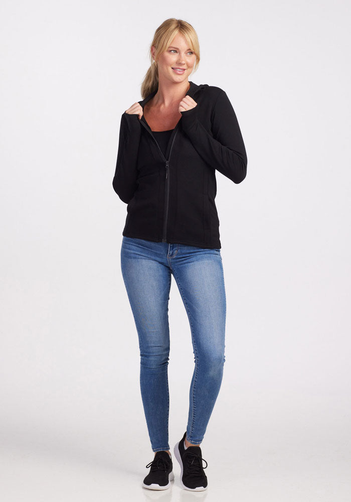 A person stands against a plain background, wearing a black Zoey Zip Up Hoodie from Woolx made with DuraLite® Fabric, blue jeans, and black athletic shoes. They are smiling and looking slightly to the side, with hands holding the hoodie near the collar. Their blonde hair is tied back.