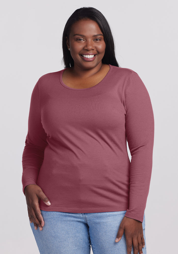 A person with long dark hair smiles while wearing the lightweight Remi Long Sleeve T-Shirt in Wild Ginger from Woolx and a pair of light blue jeans, standing against a plain, light gray background. 