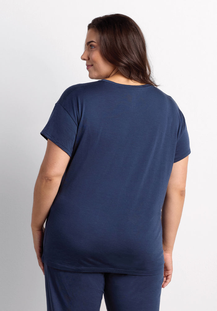 A person with long dark hair is standing and facing away, wearing a navy blue Billie Lounge Tee in the Starry Night color and matching pants from Woolx. The breathable Merino Wool fabric makes it ideal for hot sleepers. The background is plain white.