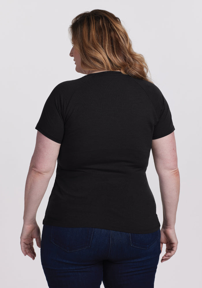 A woman with shoulder-length hair stands facing away, wearing a Woolx Mia V Neck - Black tee and dark jeans against a plain white background.