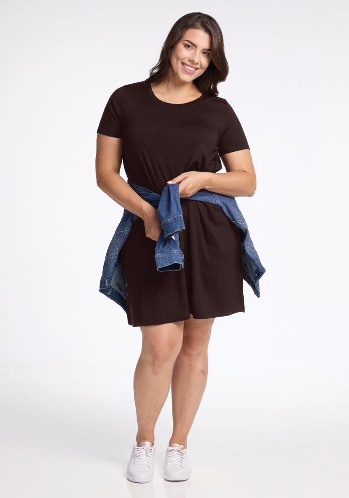 A person wearing a stylish Woolx Georgie Dress in French Roast and white sneakers stands against a plain background, smiling with a denim jacket tied around their waist.