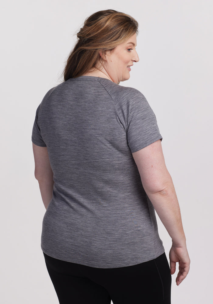 A person with long brown hair is looking to the side, wearing Woolx's Mia V Neck in Graphite Heather and dark pants. The elegant merino wool of the tee stands out against the plain white background.