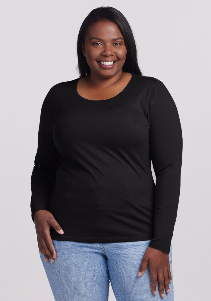 A person with long dark hair smiles warmly at the camera, wearing a Woolx Remi Long Sleeve T-Shirt in black and light blue jeans. The cozy Merino wool top complements their look as they stand against a plain light gray background. 