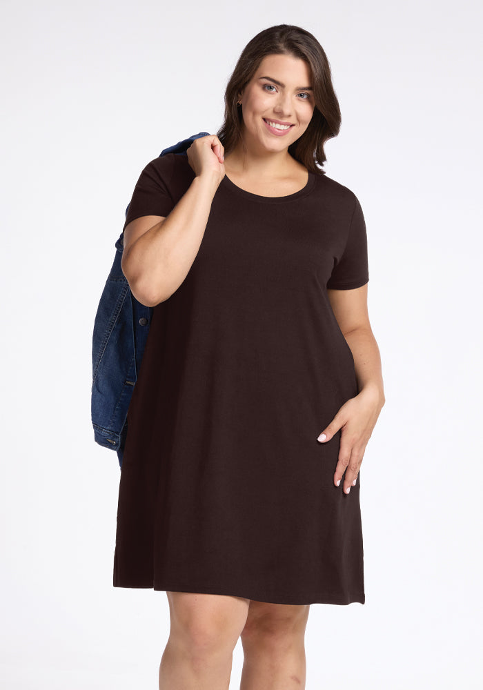 A person wearing a French Roast Georgie Dress by Woolx holds a denim jacket over one shoulder. They are smiling and standing against a plain white background. 