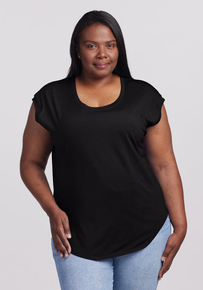 A woman with long black hair smiles, dressed in the versatile Gabi Tunic Top in black by Woolx and light blue jeans. She stands against a plain gray background, exuding effortless style. 