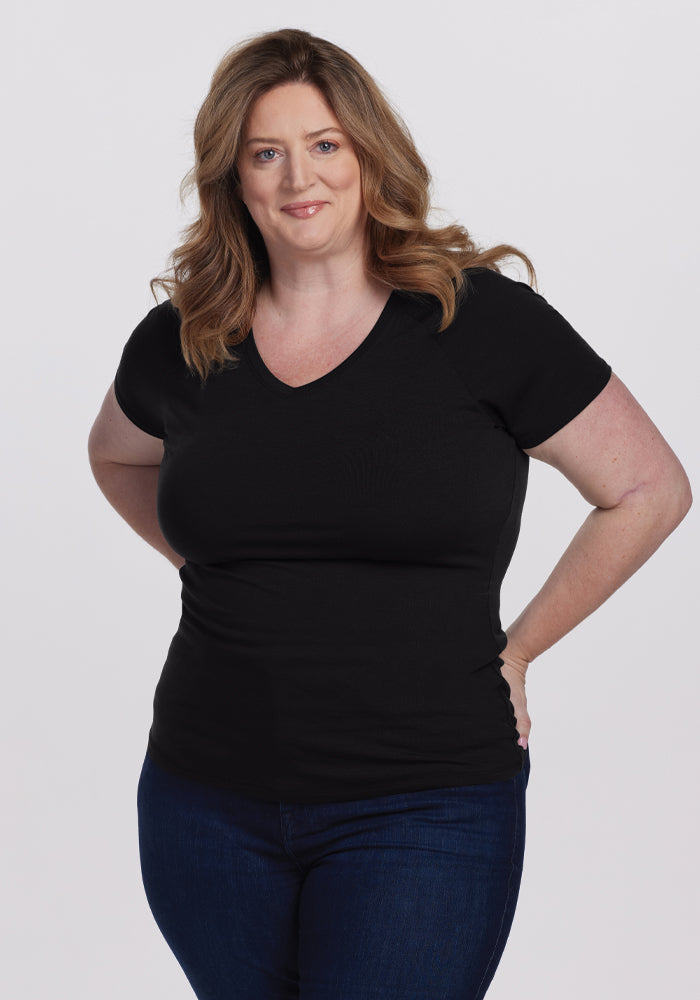 A person with long, wavy brown hair smiles slightly against a plain white background, wearing the Woolx Mia V Neck - Black in superior quality merino wool and dark jeans, hands on hips. 