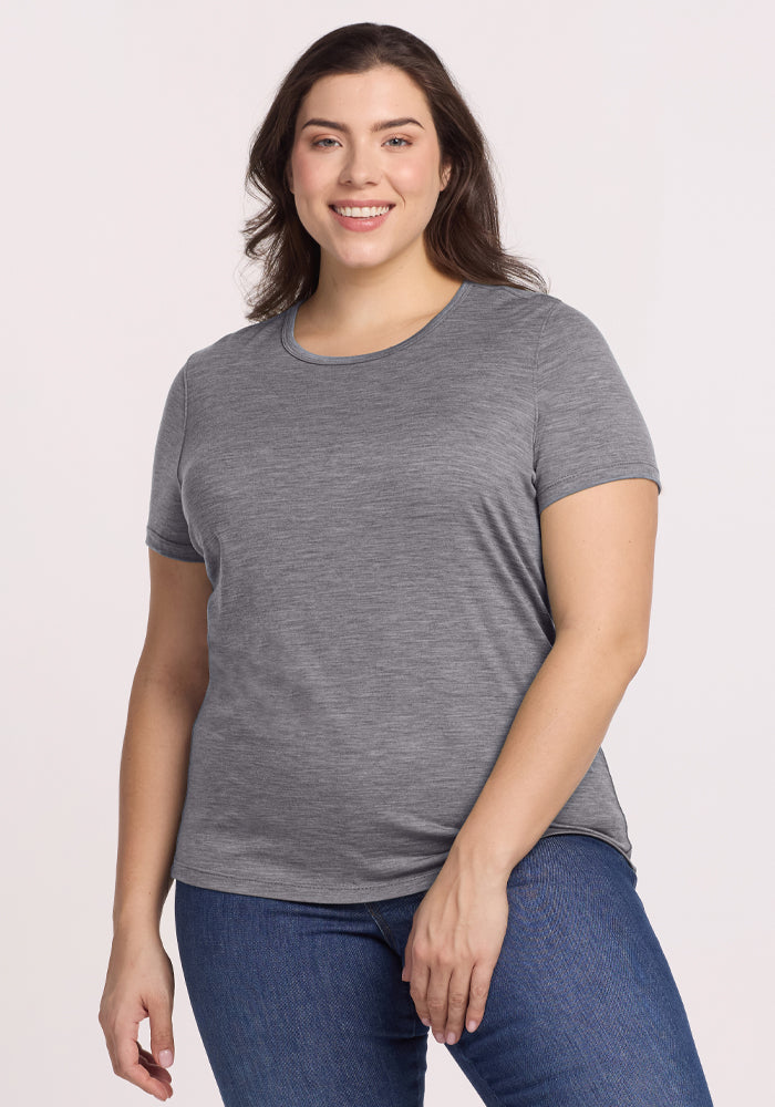 A person wearing the Woolx Liza Crew Neck Tee in Graphite Heather and blue jeans smiles while standing against a plain background. 