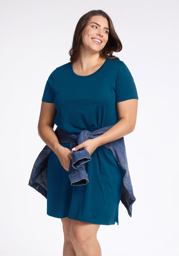 A woman wearing a short-sleeved Real Teal Georgie Dress by Woolx, with a denim jacket tied around her waist, smiles while looking to the side against a plain white background.