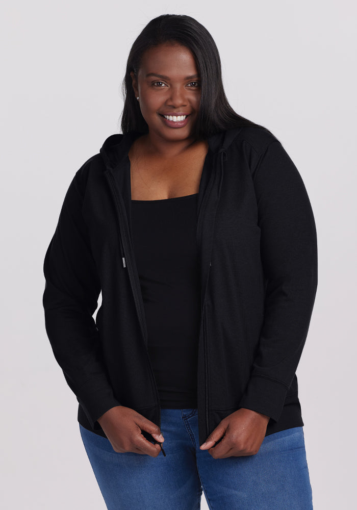 A person smiling, wearing the Woolx Ryann Hoodie in black over a black top and blue jeans, stands against a plain background. 