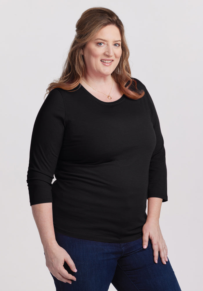 A person with long, light brown hair is smiling and posing against a plain background, wearing a versatile wardrobe staple: the Woolx Jenny 3/4 Sleeve Crew Neck in black and blue jeans, with one hand resting on their hip.