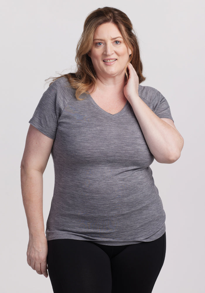 A woman with long brown hair smiles and touches her neck, wearing the comfortable Woolx Mia V Neck - Graphite Heather tee and black pants, standing against a plain white background. 