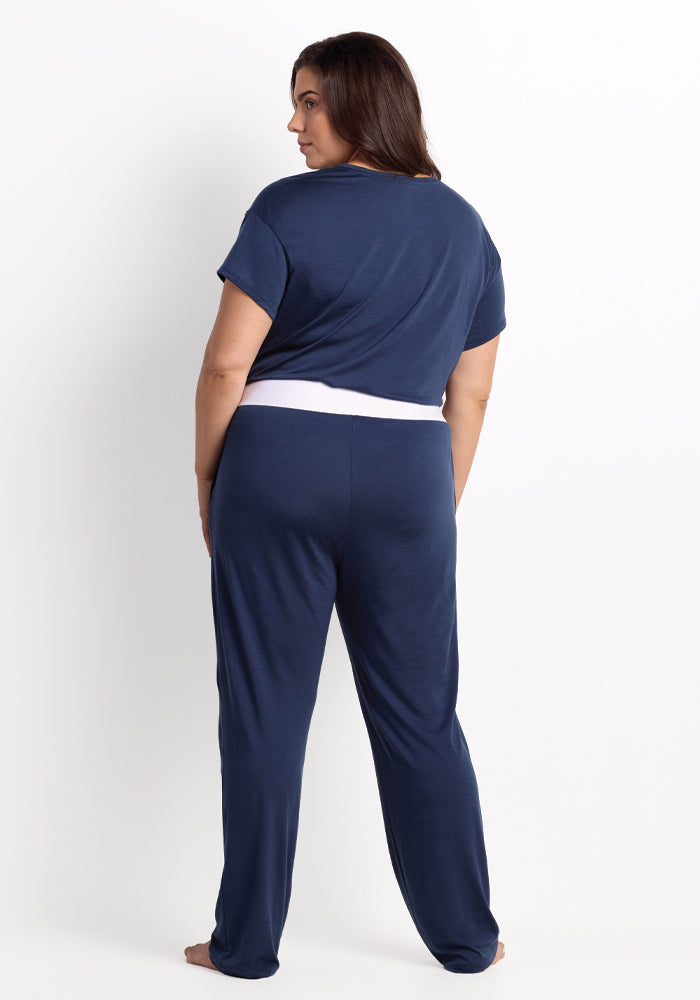 A person with long dark hair stands barefoot, facing away. They are dressed in a navy blue short-sleeve top and Maya Lounge Pants in Starry Night, featuring a white waistband. Made by Woolx from odor-resistant Merino wool, these pants offer optimal travel comfort against the plain white background.