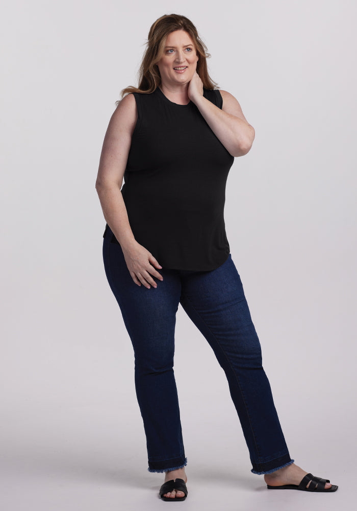 A smiling person with long hair stands against a plain background, wearing the Bella Tunic Top in black by Woolx, dark blue jeans, and black sandals. One hand is touching their neck while the other rests on their hip, showcasing relaxed summer clothing vibes.