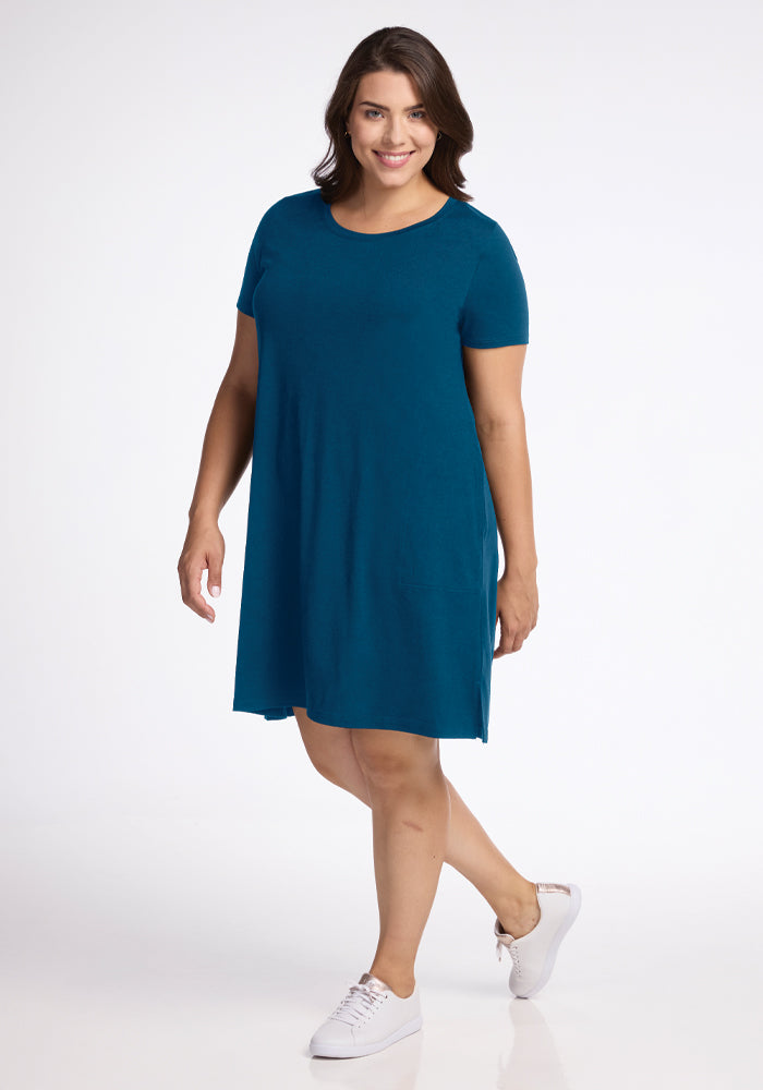 A person with long dark hair is smiling and wearing the Woolx Georgie Dress in Real Teal and white sneakers. They stand with one leg slightly raised and hand on hip against a plain white background.