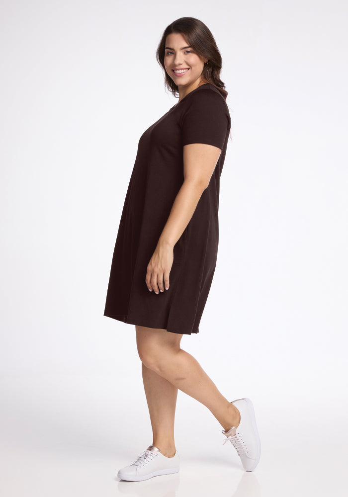 A person wearing the "Georgie Dress - French Roast" by Woolx, styled short-sleeved in a rich dark tone, pairs it with white sneakers as they pose in a studio setting. They smile while standing sideways with their left leg slightly bent forward against a plain white background.