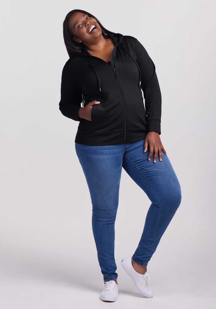 A person is smiling broadly, wearing the Woolx Ryann Hoodie in black with secure zippered pockets, blue jeans, and white sneakers. They stand against a plain gray background, slightly leaning to one side with hands in their pockets.