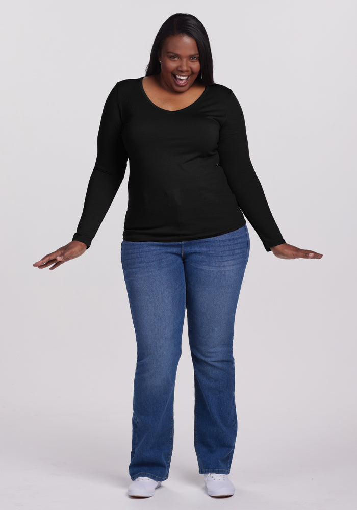 Smiling with arms slightly outstretched, a person showcases the Woolx black Layla V Neck Top. Its odor-resistant and moisture-wicking features add effortless style and functionality. Paired with blue jeans and white shoes against a plain white background, they exude casual sophistication.