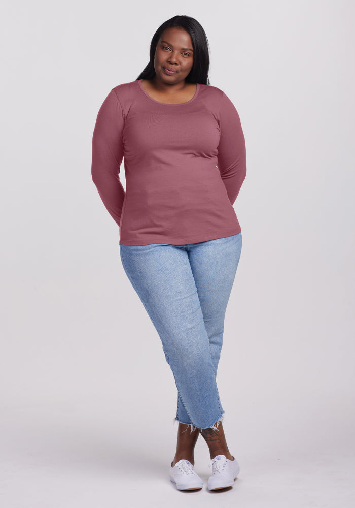 A woman poses confidently against a plain background, wearing the Remi Long Sleeve T-Shirt in Wild Ginger from Woolx, paired with light blue jeans. She stands with her legs crossed and arms relaxed, sporting white sneakers and a gentle smile.