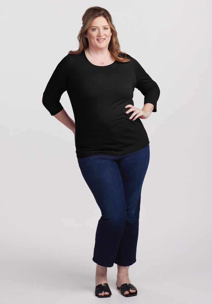 A woman with long hair stands against a white background, wearing the versatile Jenny 3/4 Sleeve Crew Neck in black merino wool by Woolx. She pairs it with dark blue jeans and black sandals, exuding a relaxed vibe, hands on her hips and smiling slightly.