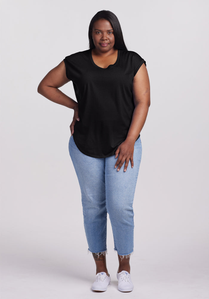 A woman wears a Woolx Gabi Tunic Top in black with light blue jeans, white sneakers, and has long, straight black hair. She stands against a plain white background with her left hand on her knee and looks at the camera neutrally.