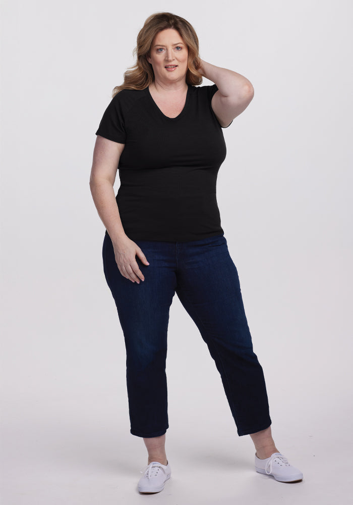 A person with long hair stands confidently against a plain background, wearing Woolx's Mia V Neck - Black Merino wool tee, dark blue jeans, and white sneakers. One hand rests on their head, exuding superior quality and effortless style.