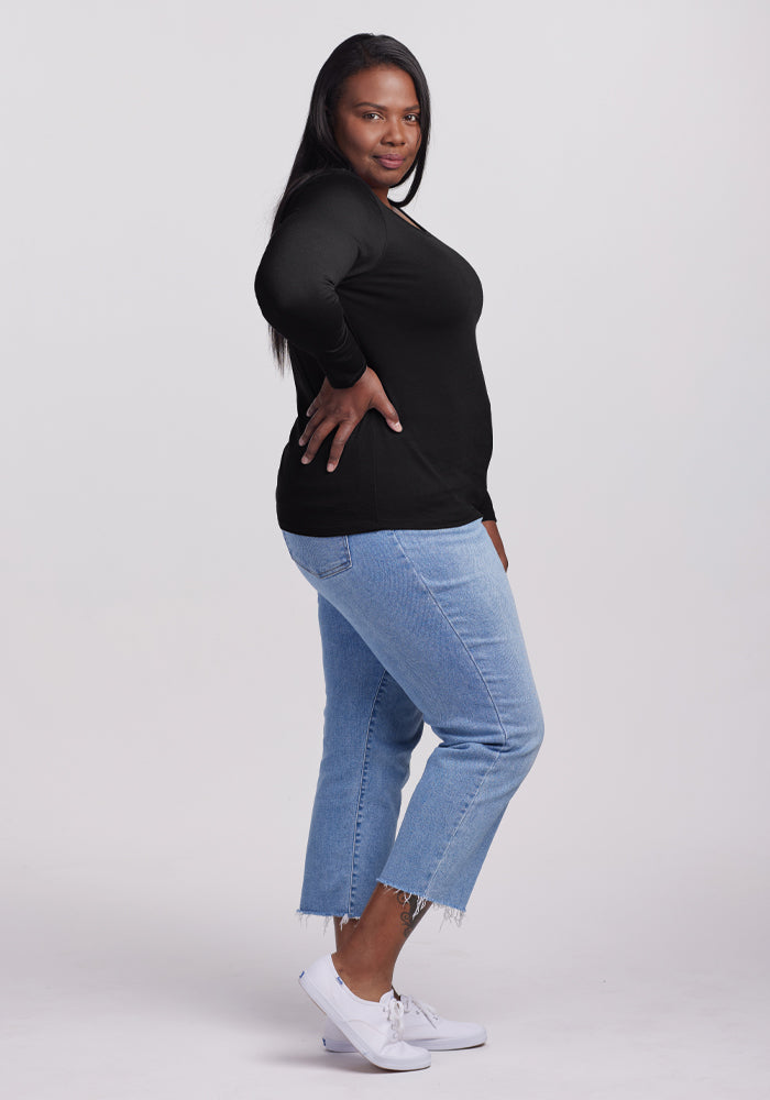 A woman with long hair stands confidently in a side pose, wearing the odor-resistant Remi Long Sleeve T-Shirt - Black by Woolx, paired with blue cropped jeans and white sneakers. She exudes casual elegance against the plain white background.