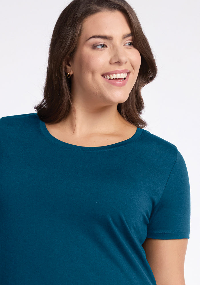 A person with shoulder-length brown hair wearing the Real Teal Georgie Dress by Woolx smiles while looking slightly to the side. The plain white backdrop highlights their casual yet chic vibe, reminiscent of a vibrant Woolx moment.