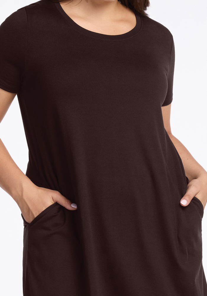 A person wearing a Woolx Georgie Dress in French Roast stands against a white background. Featuring short sleeves and pockets, the intricate details of the merino wool fabric are visible as hands rest comfortably in the pockets.