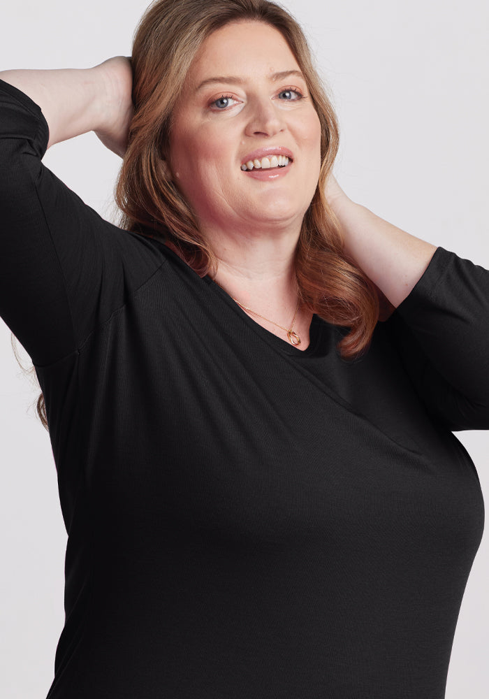 A woman with long hair smiles at the camera, exuding effortless style. She wears the Jenny 3/4 Sleeve Crew Neck in black by Woolx, a versatile wardrobe staple, with her hands behind her head. The plain, light-colored background enhances her modern elegance.