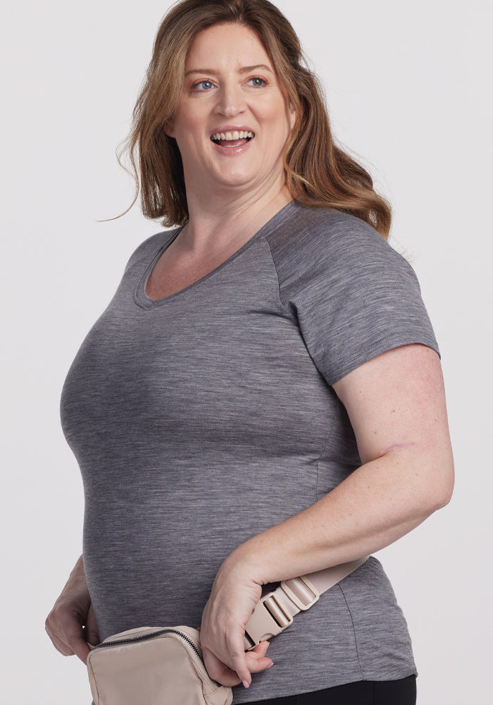 A woman with long brown hair smiles while holding a beige bag and wearing the Woolx Mia V Neck in Graphite Heather. She stands against a plain white background.