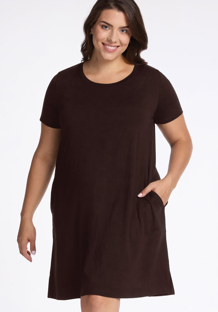 A woman wearing a short-sleeved, Georgie Dress in French Roast by Woolx stands smiling, with one hand in the dress pocket. The knee-length A-line style beautifully complements her dark hair against a plain white background.